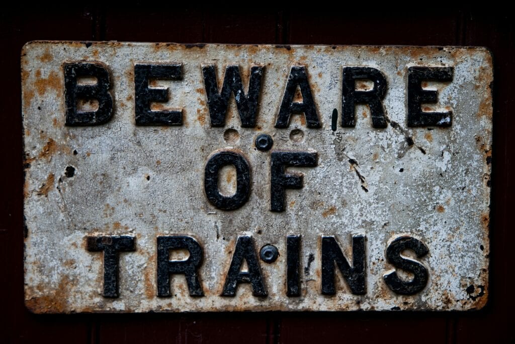 A weathered metal sign with bold black letters reads "BEWARE OF TRAINS" against a dark background.