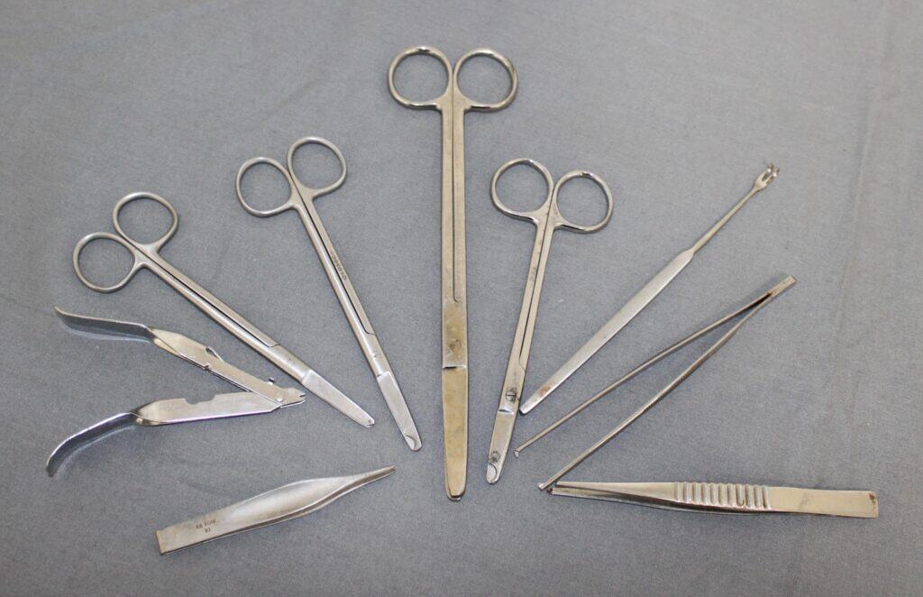A set of various metal surgical instruments, including scissors, forceps, tweezers, and other tools, arranged in a semi-circle on a grey surface.
