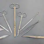 A set of various metal surgical instruments, including scissors, forceps, tweezers, and other tools, arranged in a semi-circle on a grey surface.
